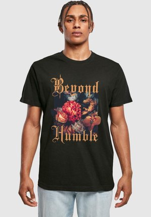 Sort bomulds t-shirt med et blomstermønster i klare farver og guldskrift, der siger "Beyond Humble." Enkel crew neck design.