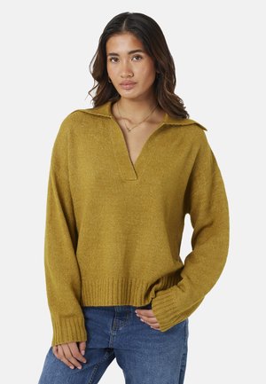 Frau mit langen dunklen Haaren, die einen senfgelben Pullover mit Kragen und blaue Jeans trägt, steht vor einem schlichten weißen Hintergrund.