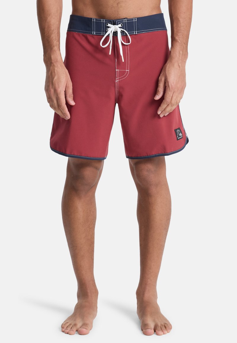 Červené plavky s tmavomodrým pásom, bielym sťahovacím šnúrkou a kontrastným prešívaním. Na spodnej ľavej strane je logo záplata.