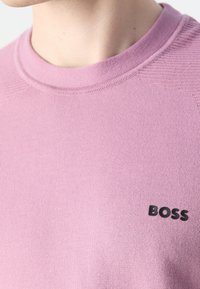 Gros plan sur une personne portant un pull en maille violet clair avec un logo noir "BOSS" sur la poitrine.