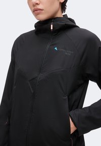 Άτομο που φοράει μαύρο μπουφάν softshell με ψηλό γιακά, φερμουάρ μπροστά και ένα μικρό μπλε λογότυπο στο στήθος, με το χέρι στην τσέπη.