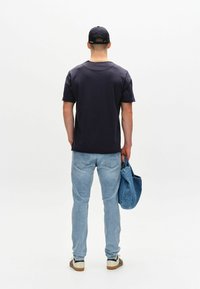 Mornarska kratka majica s kratkimi rokavi, svetlo modri kavbojke, drži modro denim torbo, stoji obrnjen proti proč, nosi črno kapo. Preprost in sproščen videz.