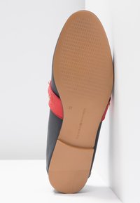 Chaussure noire à enfiler avec une lanière rouge, semelle plate, dessous en caoutchouc texturé et un accent doré près de la lanière. Taille 37, logo de la marque visible.