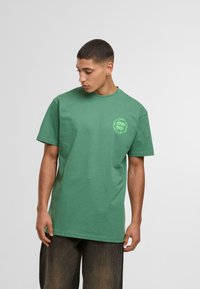 Camiseta de algodón verde con cuello redondo y mangas cortas. Presenta un logo verde neón en el lado izquierdo del pecho. Textura suave y corte relajado.