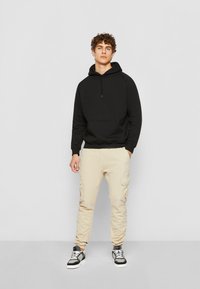 Svart hettegenser med snorer, frontlomme; parret med beige cargo-bukser med sidelommer; svart og hvit treningsko.