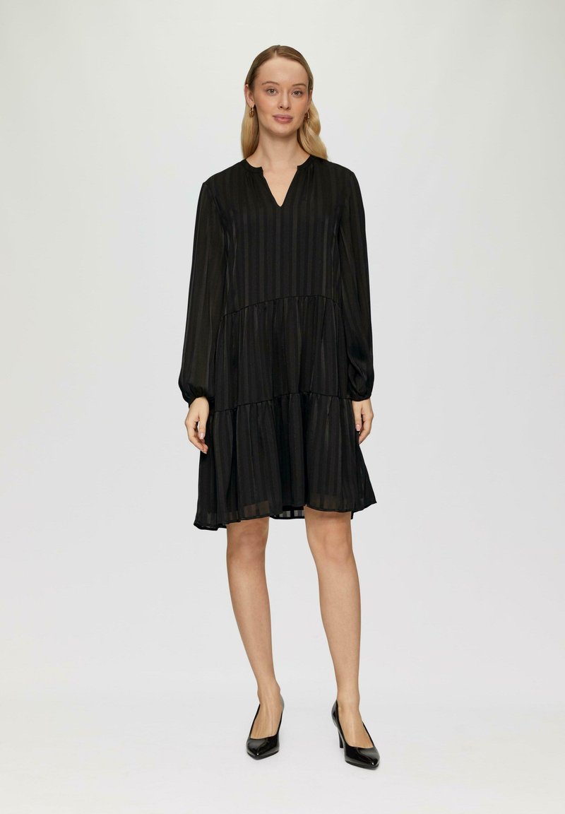 Robe noire à volants avec manches longues, décolleté en V et rayures verticales. Tissu léger et fluide avec un éclat subtil, associée à des talons noirs.