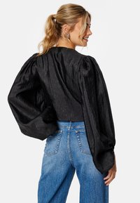 Zwarte, textuurblouse met lange, opgeblazen mouwen, gecombineerd met hooggetailleerde blauwe jeans. Heeft een ronde halslijn en een detail op de rug.