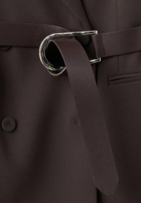 Brązowy materiałowy pasek z dużą srebrną okrągłą klamrą, przypięty do ciemnobrązowej marynarki z podwójnym rzędem guzików i widocznymi guzikami.