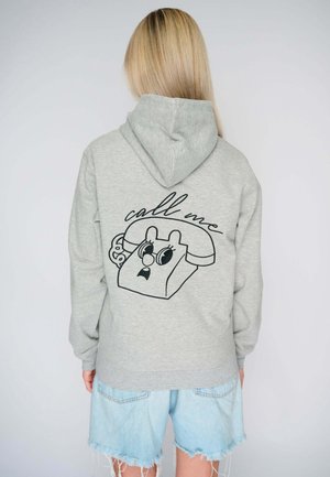 Mujer con cabello largo y rubio, que lleva una sudadera gris con capucha con un dibujo de un teléfono y el texto "llámame" en la parte posterior, combinado con pantalones cortos de mezclilla azul clara.