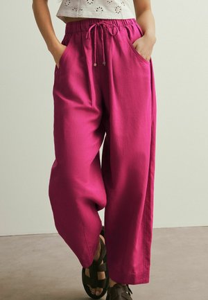 Mujer con pantalones magenta holgados con cordón y bolsillos, y sandalias negras, de pie con las manos en los bolsillos contra un fondo liso.