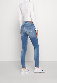 Jeans ajustados azules con cintura alta, parte frontal desgastada, dos bolsillos traseros y una etiqueta de cuero con marca. Combinados con zapatillas blancas y una camiseta ajustada.