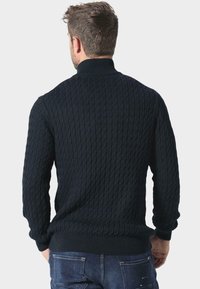 Ciemnoniebieski sweter o dzianinowej strukturze z wysokim kołnierzem, wyróżniający się fakturowanym wzorem w warkocze oraz ściągaczami przy mankietach i dolnej krawędzi. Noszony z dżinsowymi spodniami.