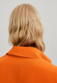Orange collared shirt with a smooth texture, worn from the back. Hair has a wavy style, with a light blonde color.