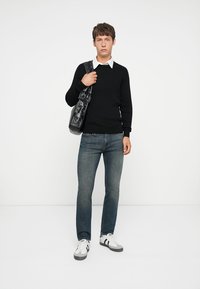 Pulover negru cu textură, cămașă albă cu guler dedesubt, blugi, și geantă neagră de umăr. Modelul poartă adidași albi cu accente negre.