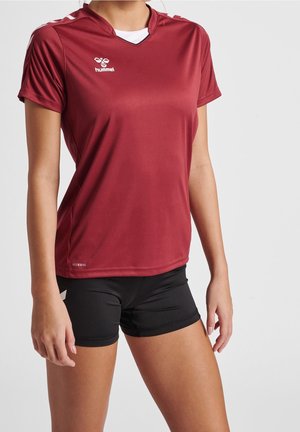Vrouw draagt een bordeauxrood sportshirt met korte mouwen en zwarte sportkorte broek, staand tegen een effen lichte achtergrond.
