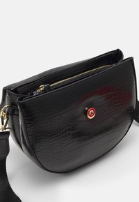 Schwarze Umhängetasche aus Leder im Krokodil-Design mit runder Form, goldenen Beschlägen und einem auffälligen roten Akzentknopf. Mit Reißverschlussfach.