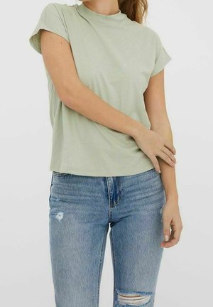 Vrouw met lichtgroen T-shirt met korte mouwen en blauwe gescheurde spijkerbroek, staand met één hand die de tegenovergestelde arm aanraakt tegen een effen achtergrond.