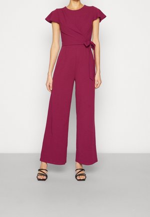 Kvinde iført en ærmeløs magenta jumpsuit med vide ben og et bindebånd i taljen, kombineret med sorte sandaler med hæl og stropper.