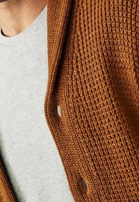 Cardigan lavorato a maglia marrone con texture, dotato di apertura frontale, caratterizzato da una trama allentata e fori per i bottoni, indossato sopra una camicia grigia chiaro.