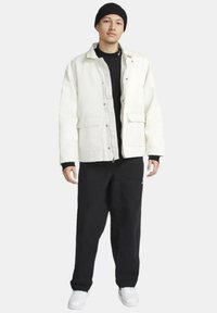 Chaqueta blanca de lona con cremallera frontal y bolsillos, llevada sobre una camiseta negra, combinada con pantalones negros y zapatillas blancas. Gorra negra en la cabeza.