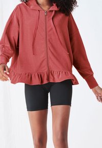 Sweat à zip couleur rouille avec un ourlet à volants, des manches longues et une poche avant. Assorti avec un short de vélo noir. Texture lisse.