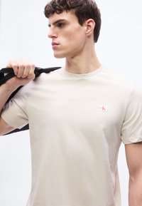 Νεαρός άνδρας που φοράει μπεζ t-shirt Calvin Klein, κρατώντας ένα μαύρο μπουφάν στον ώμο του, κοιτάζοντας προς τα πλάγια με φόντο ένα λευκό χρώμα.