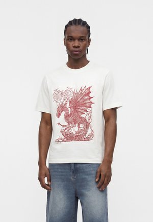 Mladý muž s vrkočmi, ktorý nosí biele tričko s detailnou ilustráciou červeného draka, v kombinácii s vybledlými modrými džínami.