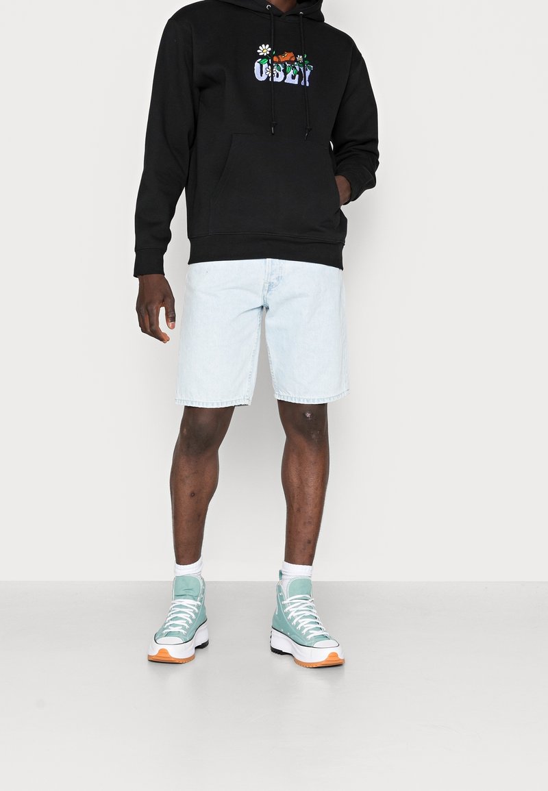 Svart hoodie med blommigt "OBEY" mönster, kombinerad med ljusblå denimshorts och turkosa högskurna sneakers med orange detaljer.