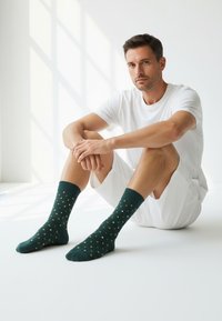 Zelene nogavice z majhnimi rumenimi in belimi trikotnimi vzorci, mešanica bombaža, dolžine do gležnjev, nosi jih oseba, ki sedi v beli okolici.