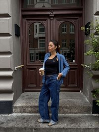 Modele melnā tanktopā un gaišās džinsa pogātās krekla virskrekliņā, ģērbta plašās zilās džinsu biksēs ar kabatu, tur dzērienu caurspīdīgā glāzē.