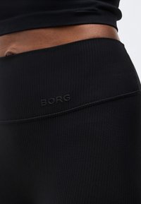 Κοντινή λήψη της μέσης ενός ατόμου που φοράει μαύρο ριμπ κολάν με ψηλή μέση και την επιγραφή "BORG" ανάγλυφη στην μπροστινή αριστερή πλευρά.