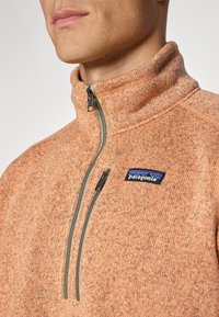Pullover arancione realizzato in tessuto testurizzato con colletto alto, chiusura con zip e una toppa del logo Patagonia blu sul lato sinistro.