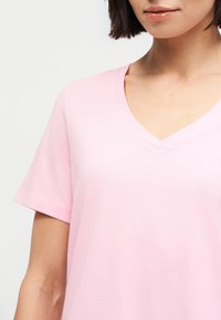 Γυναίκα που φοράει ανοιχτό ροζ T-shirt με Vλαιμόκοψη και κοντά μανίκια, με απλό φόντο, εμφανίζοντας τον λαιμό, τα χείλη και ένα μέρος του βραχίονα της.