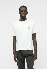 Camiseta blanca de algodón de manga corta, con un pequeño logo circular de color óxido en el pecho. Cuello redondeado simple, corte relajado.