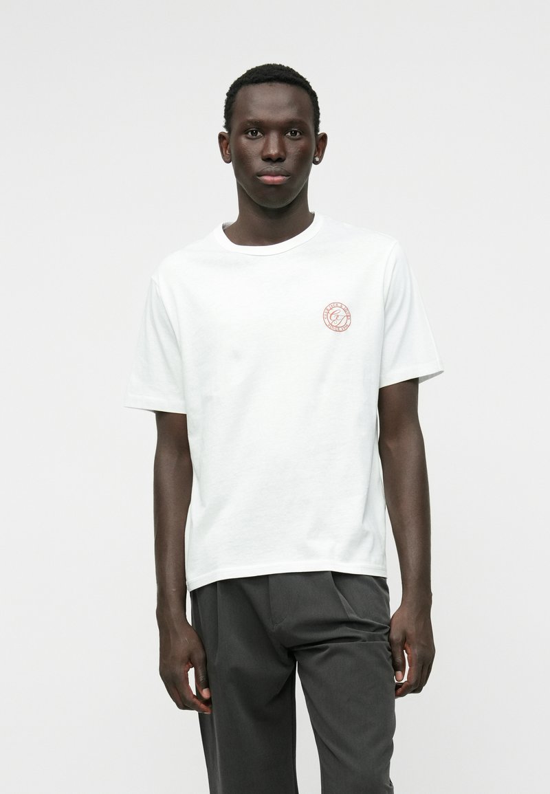 Camiseta blanca de algodón de manga corta, con un pequeño logo circular de color óxido en el pecho. Cuello redondeado simple, corte relajado.