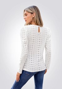 Witte blouse met lange mouwen, voorzien van een textuur met bloemenpatroon en een sleutelgatdetail aan de achterkant, gecombineerd met blauwe spijkerbroeken. Soepele stof, aansluitend ontwerp.