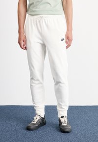 Fatos de treino brancos com um cós elástico e logótipo preto da Nike, ajustados nos tornozelos. Combinados com sapatilhas pretas e atacadores cremosos.