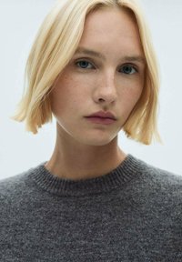 Blonde lase s plošno šišanjem okvirja obraz z madeži, oblečen v temno sivo teksturirano pulover s krojnim izrezom.
