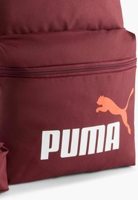 Bordeauxkleurige stoffen rugzak met een voorzak met ritssluiting, een opvallend wit "PUMA" logo en een oranje springende poema afbeelding.