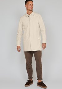 Abrigo impermeable de color beige claro, con cuello, cierre de un solo botón y bolsillos laterales. Combinado con pantalones marrones y zapatos oscuros.