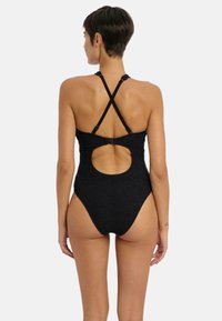 Maillot de bain une pièce noir avec une surface texturée, des bretelles croisées et une découpes circulaire dans le dos. Les ouvertures de jambe à haute découpe mettent en valeur la silhouette.