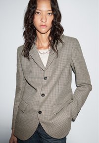 Geruite blazer in bruin en crème met pieklapels, drie knopen en functionele zakken. Gedragen over een lichtgekleurde top.