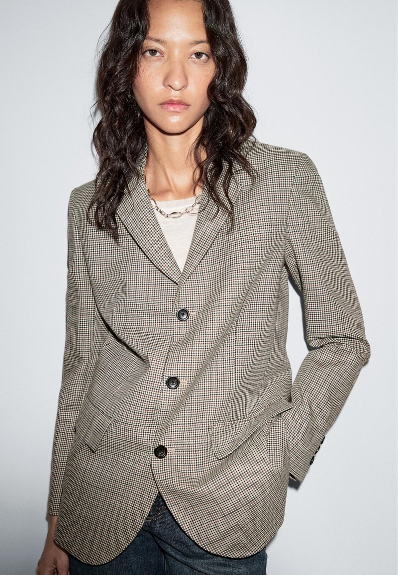 Geruite blazer in bruin en crème met pieklapels, drie knopen en functionele zakken. Gedragen over een lichtgekleurde top.