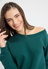Sudadera verde sin hombros con mangas largas, textura suave y diseño sencillo. Modelo con cabello castaño claro sonríe, con la mano cerca de la cara.