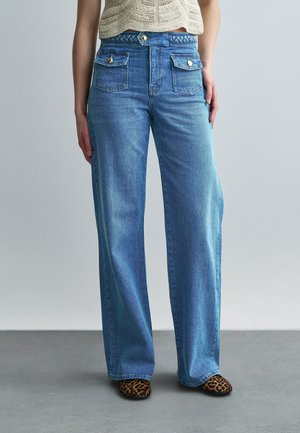 Žena nosi svetlomodré široké rifle s vysokým pásom, predné vrecká na gombíky, pletený top a topánky so vzorom leopardích škvŕn.