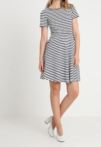 Žena v šatech s krátkým rukávem, černobílých pruhovaných, s bílými podpatky, stojící na neutrálním světle pozadí.