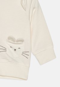 Cream-colored long-sleeve shirt made of soft fabric, featuring a pocket with a rabbit face design and stitched whiskers.