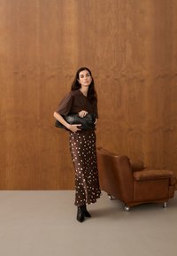 Femme debout à côté d'un fauteuil en cuir marron, portant une jupe marron à pois, un haut sombre, des bottes noires, tenant une grande pochette noire.