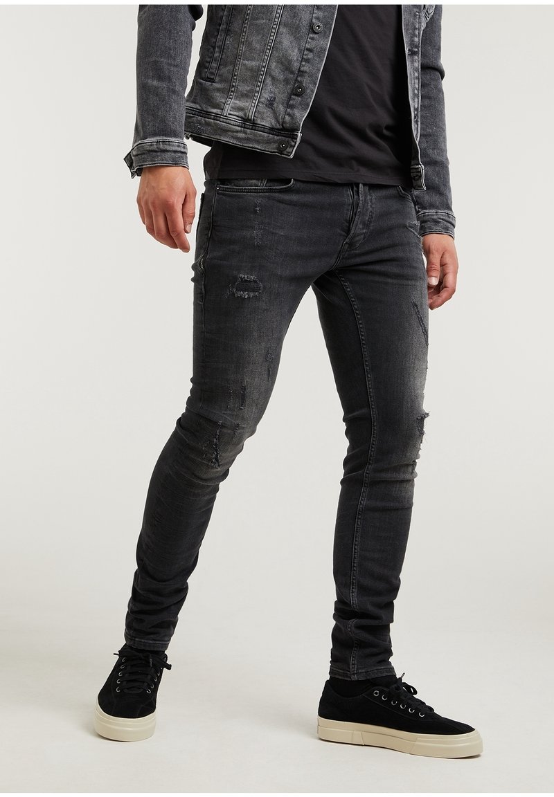 Homme portant un jean skinny noir déchiré, des baskets noires avec des semelles blanches, et une veste en jean noire par-dessus une chemise noire.