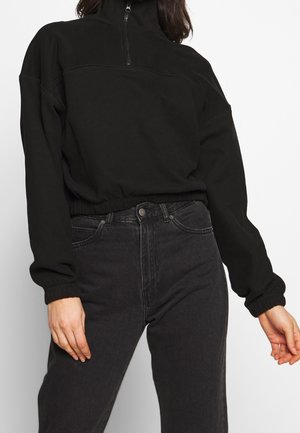 Svart kortad sweatshirt med hög krage, halvdragkedja och elastiska muddar. Kombinerad med högmidjade svarta jeans.